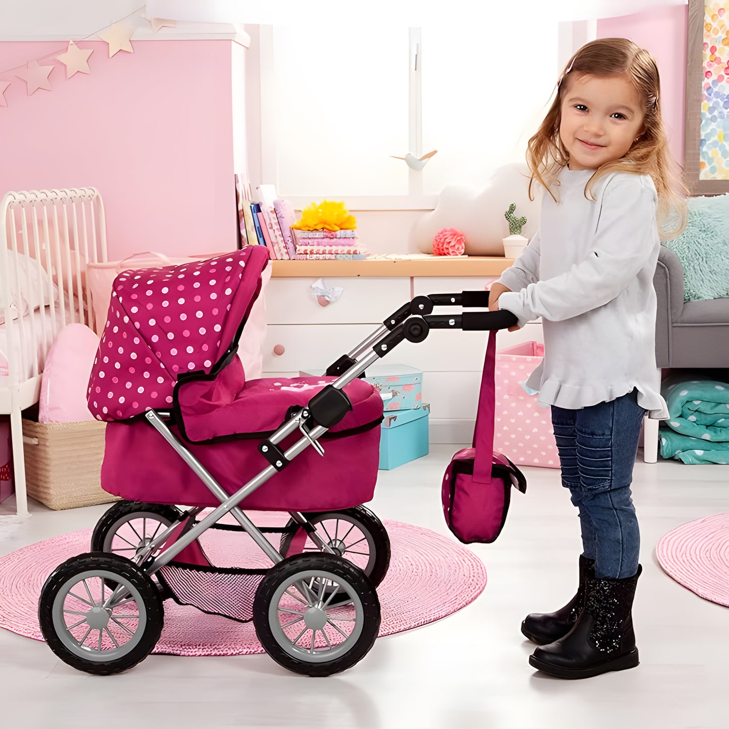 A young girl with blonde hair stands next to a pink stroller toy in a pink children's bedroom. The stroller has a pink fabric body with white polka dots on the canopy, a silver metal frame, and black wheels.