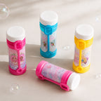 Four bubble bottles with white caps arranged on a light beige surface with floating soap bubbles. Three bottles stand upright, two are pink and one is blue, and one bottle lies on its side.